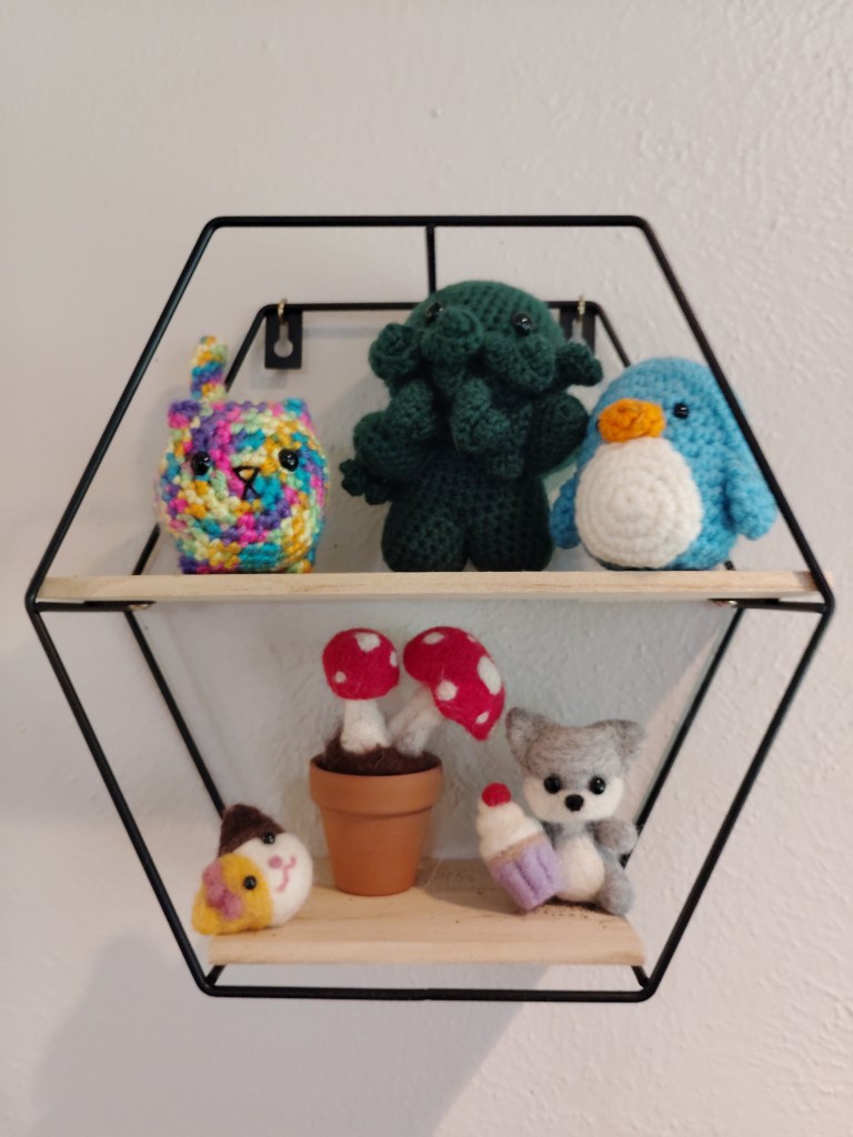 Several crafting projects sit on a small hexagonal shelf. Top: A multicolored cat loaf, green cthulu, and blue bird amigurumi. Bottom: Needle felted tri-colored cat head charm, red and white mushrooms in a clay pot, and a gray and white wolf holding a cupcake with a purple liner and red cherry on top