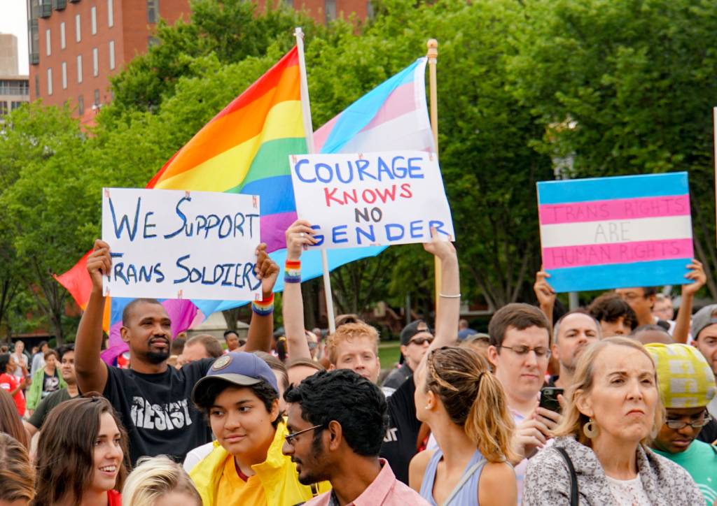 Photo of a group of protestors waving gay and trans pride flags. Two signs are being held up by two men: "We support trans soldiers: and "Courage knows no gender". 
From the Stop Transgender Military Ban in Washington, DC. on 07-29-2014